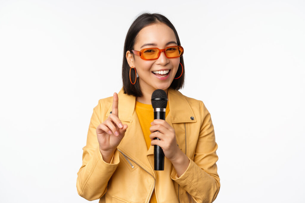 Người phụ nữ châu Á xinh đẹp với kính râm, cô gái sành điệu hát, phát biểu bằng micrô, cầm micrô, mỉm cười, đứng trên nền trắng với không gian bản sao thể hiện sự tự tin và khả năng giao tiếp của một host chuyên nghiệp